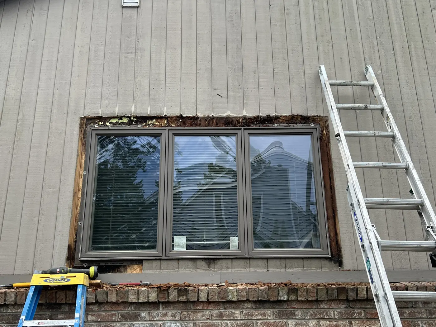 An exterior window with the surrounding wood stripped back for replacement, a ladder resting alongside.